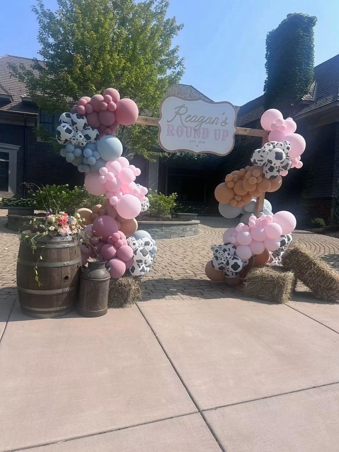 A balloon arch flanked by hay bales and a wooden barrel stands over a sign reading 'Reagan's Round Up, Est. 2018' on a paved driveway.