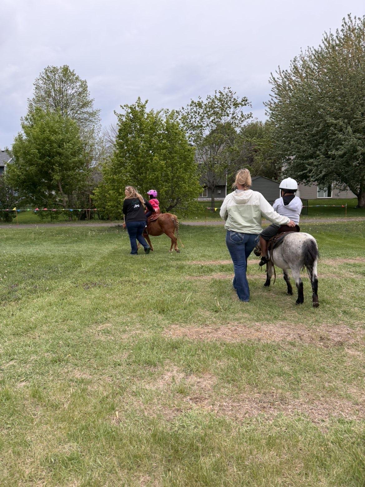 A man leads a pony carrying a girl in a fringed vest and floral cowboy hat across a backyard lawn, with a pool visible in the distance.