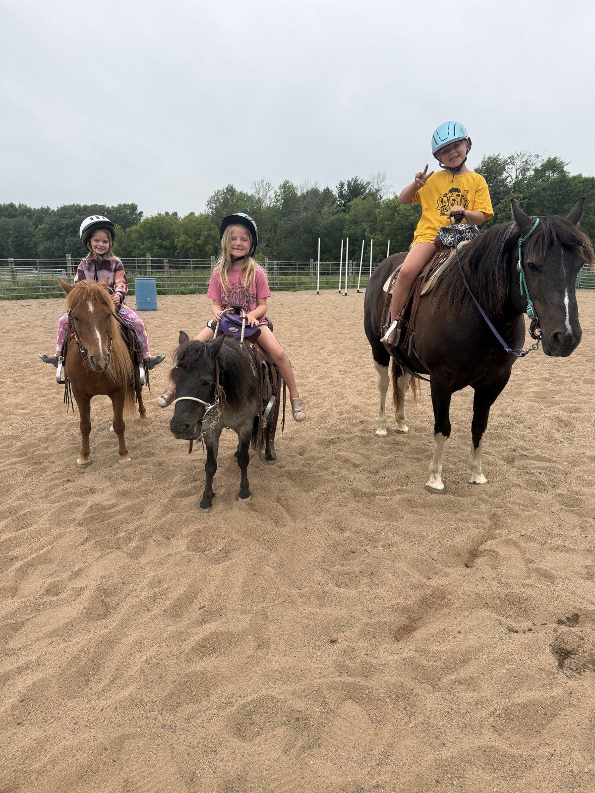 Three children in riding helmets sit on ponies side by side in a sand arena.