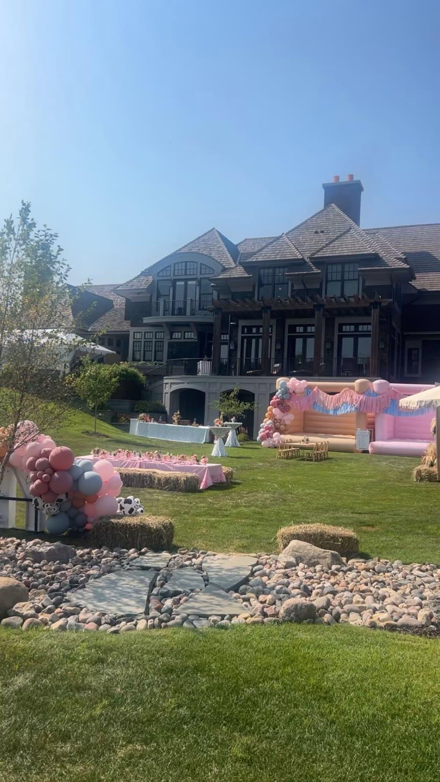 A large home's backyard set up with balloon garlands, hay bale seating, tables, and inflatable bounce houses; no animals are visible in frame.