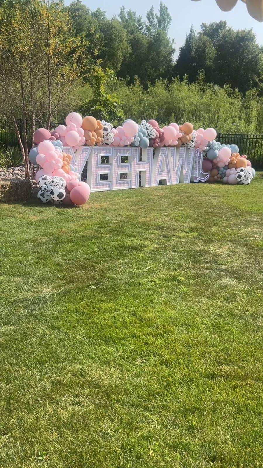 A balloon garland spelling 'YEEHAW' with cow-print balloon accents stands on a lawn in front of trees; no people or animals visible.