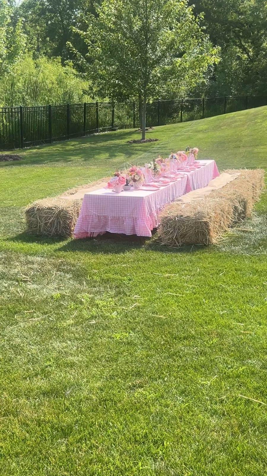 A long table with a pink gingham cloth and floral centerpieces sits between two hay bales on a lawn near a fence line.