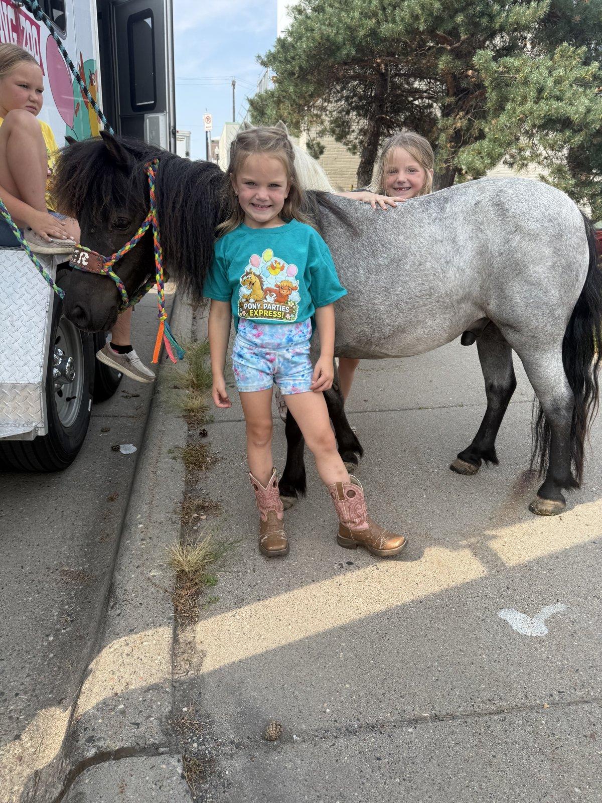 Two girls pet a gray pony and a dark pony beside the Pony Parties Express trailer.