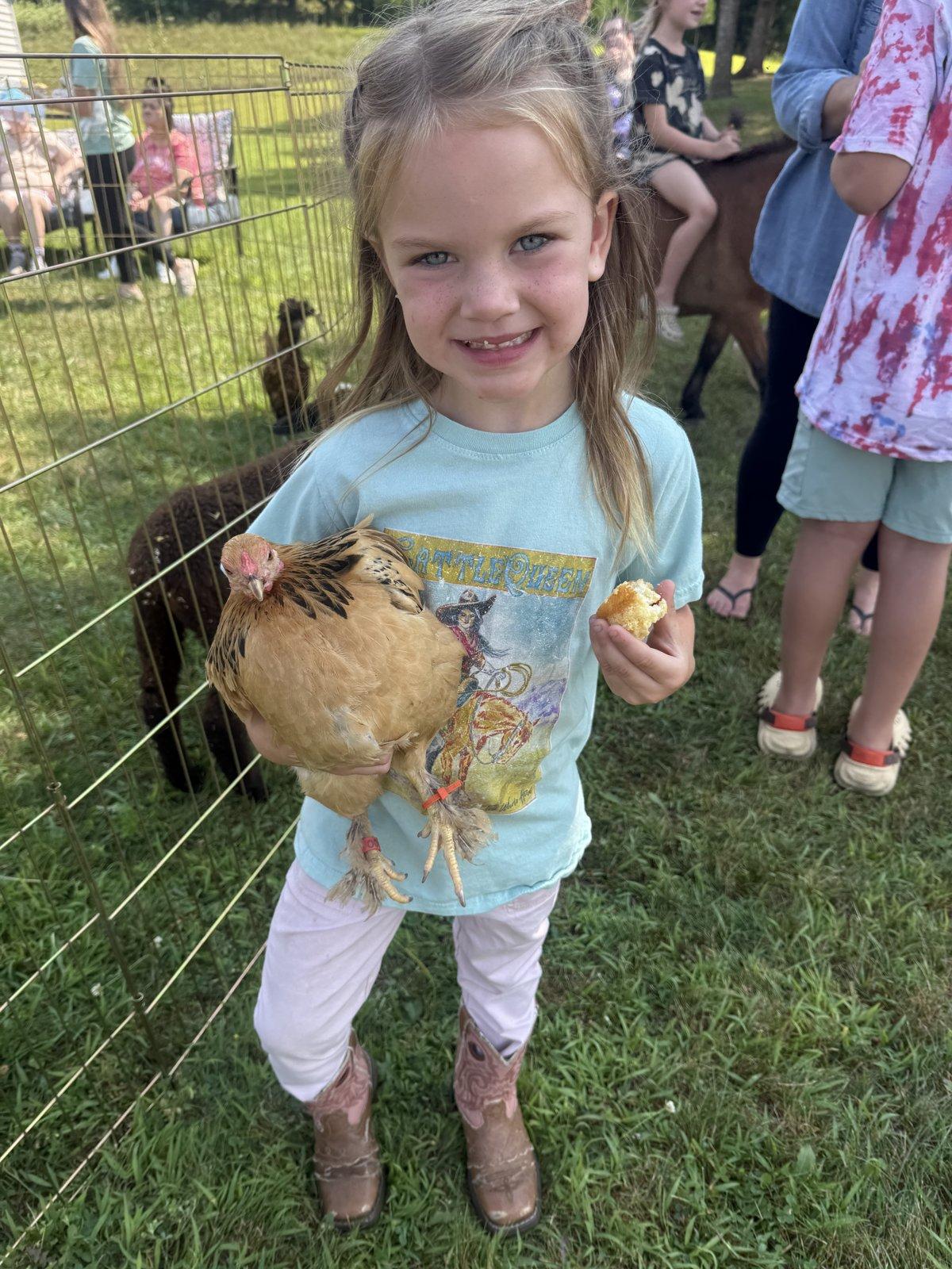 A girl in cowgirl boots holds a chicken beside a petting pen in a grassy yard.