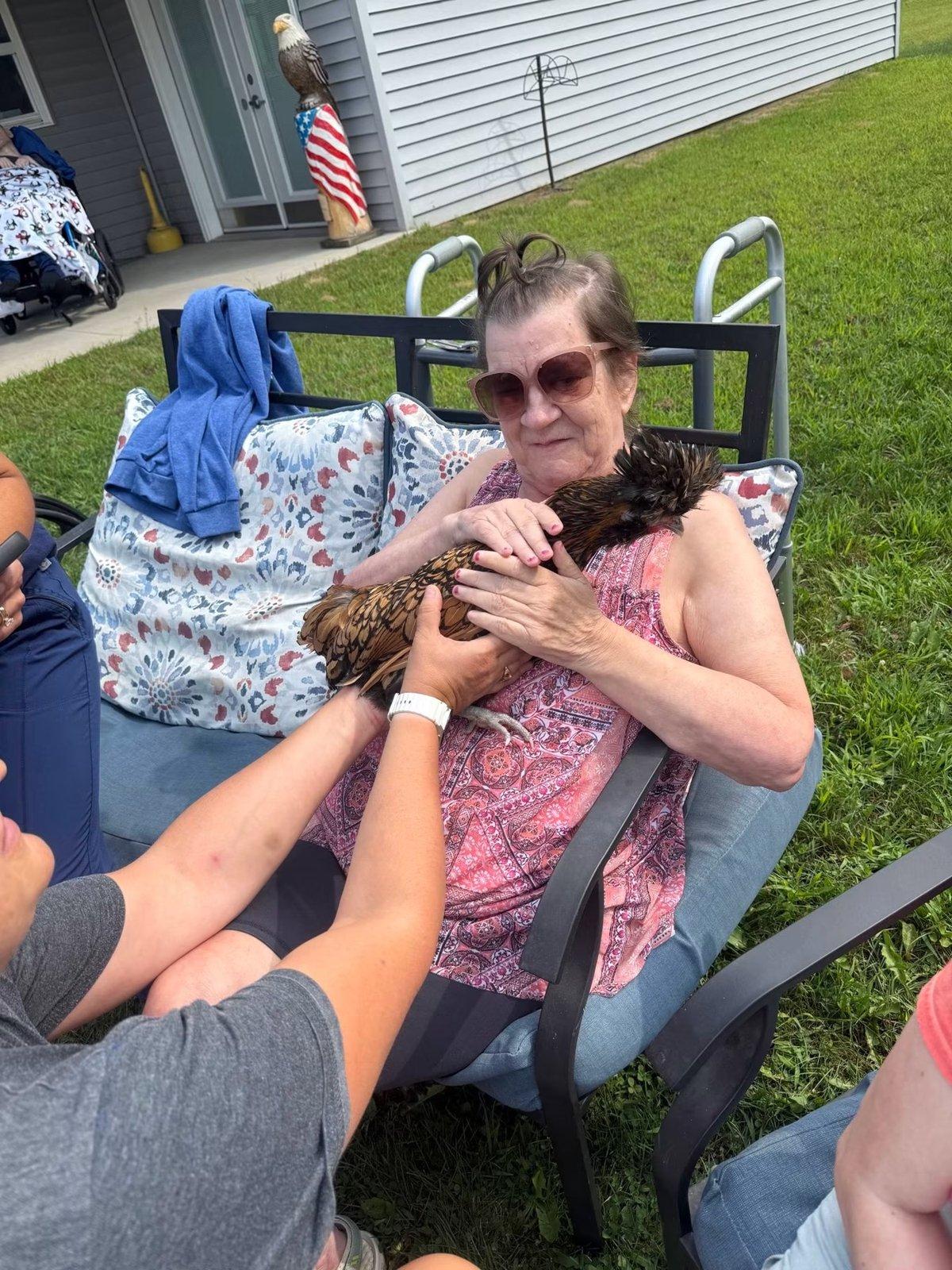A seated woman holds a chicken with a helping hand from a caregiver.