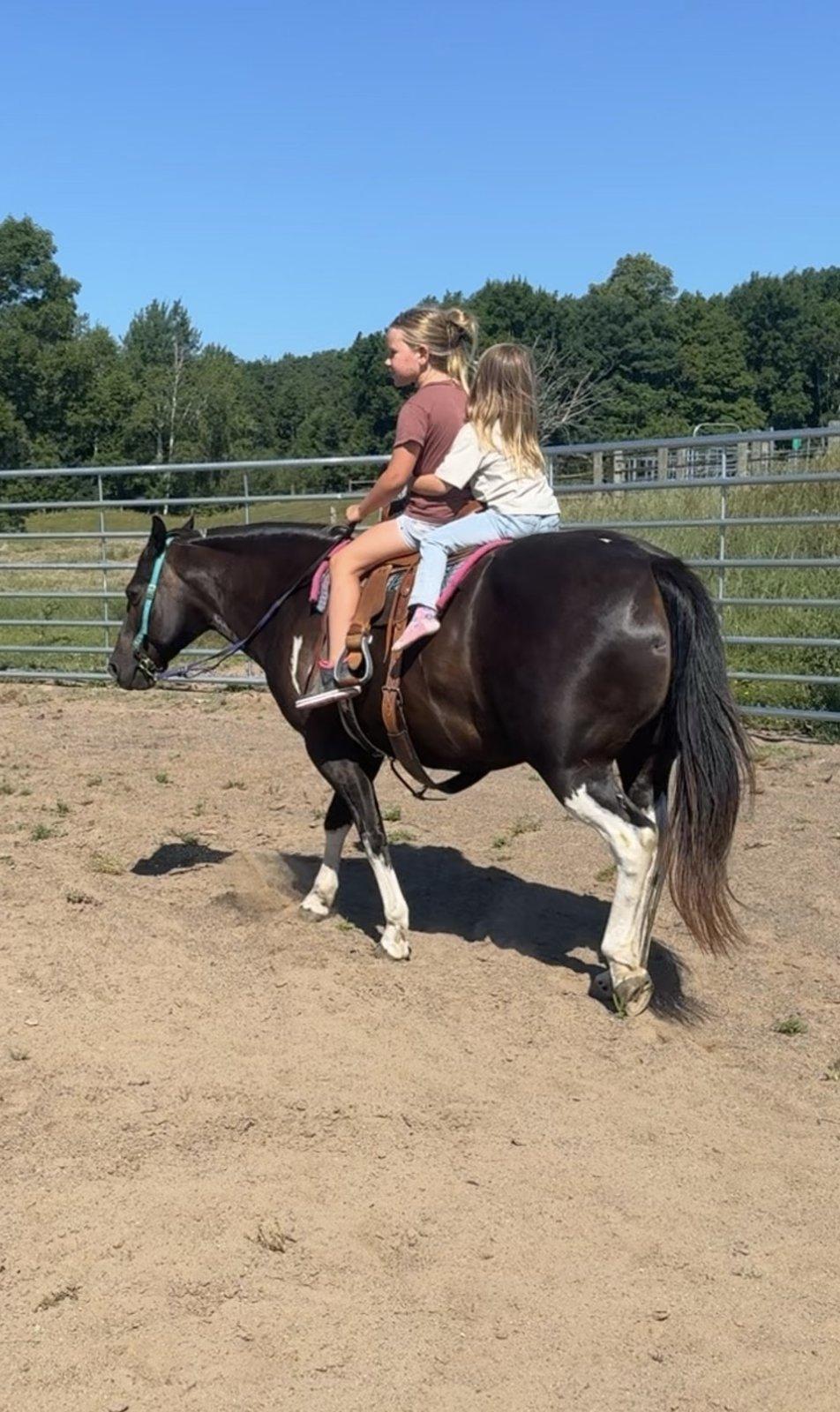 Two girls ride together on a black-and-white pinto pony inside a fenced sand arena.