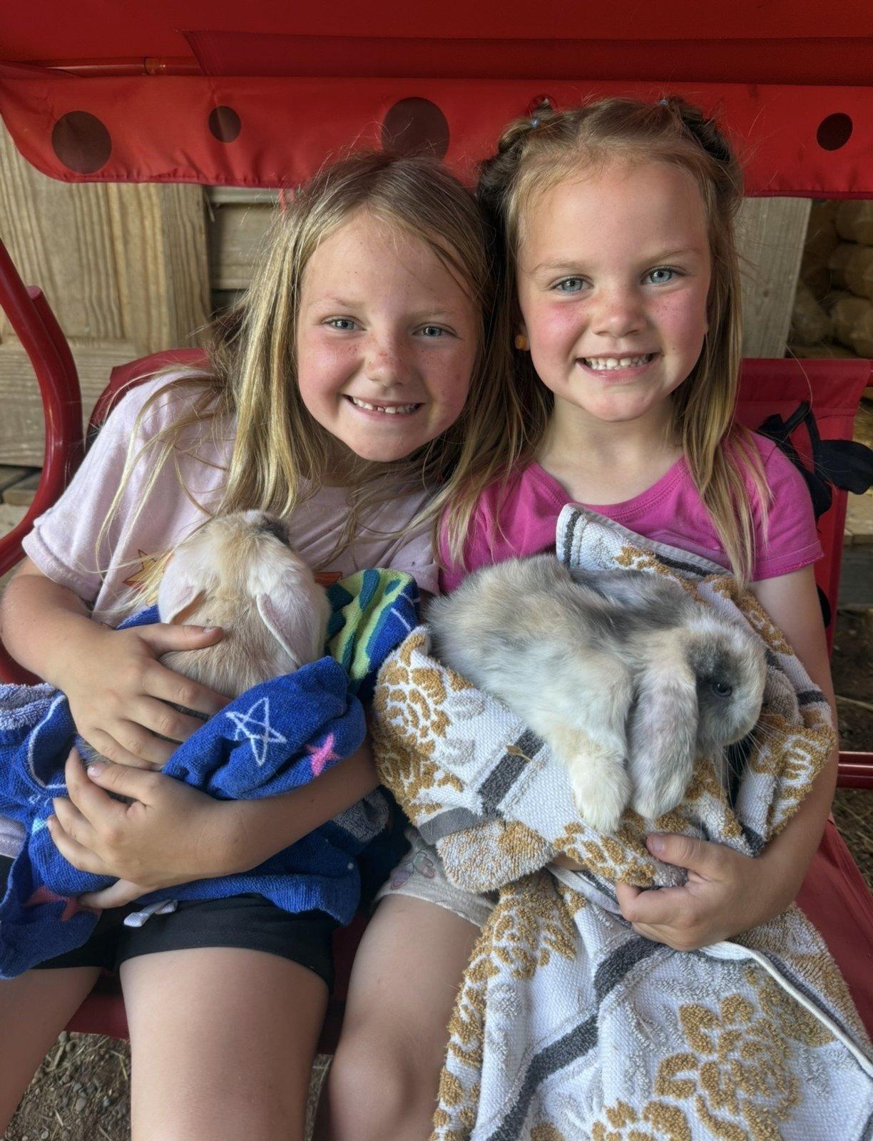 Two girls sit on a porch swing, each holding a lop-eared rabbit wrapped in a towel.