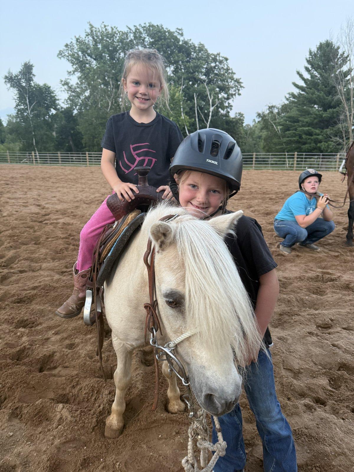 Two girls stand beside a cream-colored pony in a sand riding arena.