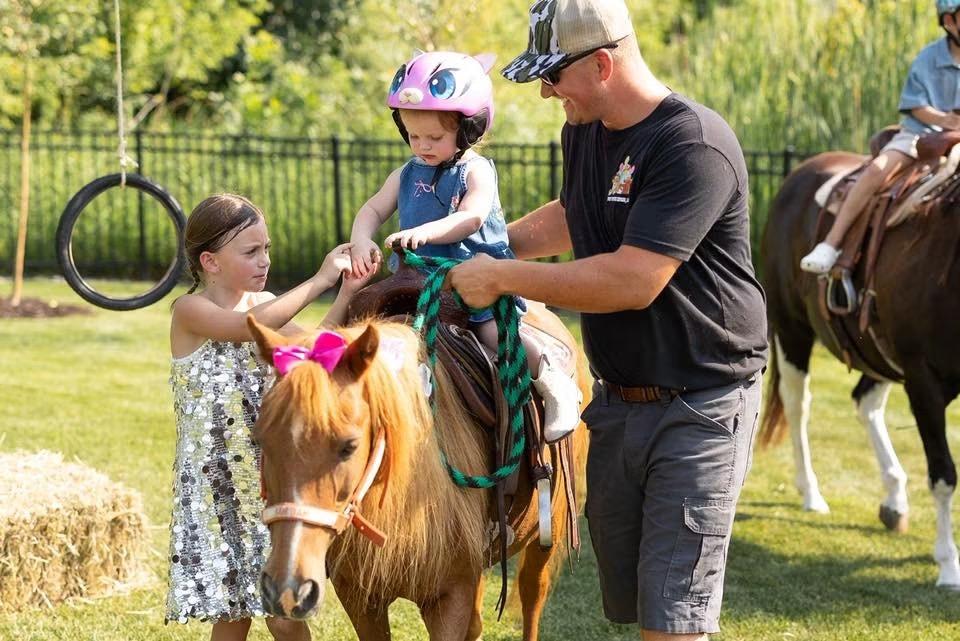 A man helps a small child in a helmet sit on a pony while another girl looks on, with a second pony and rider behind them, near a tire swing and hay bale.