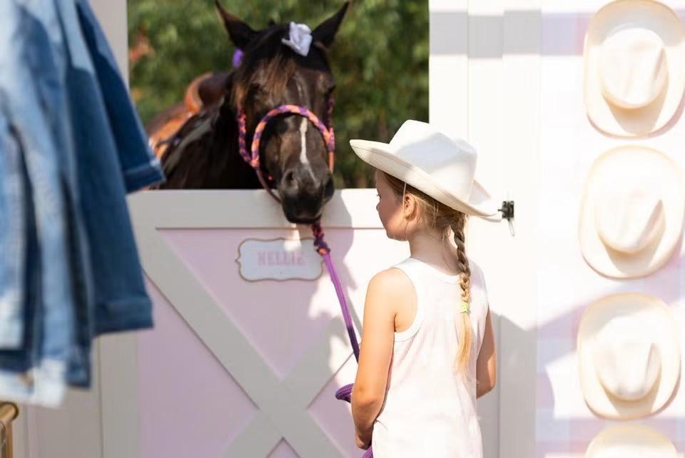 A girl in a white hat stands facing a horse looking over a decorated stall door labeled Nellie.