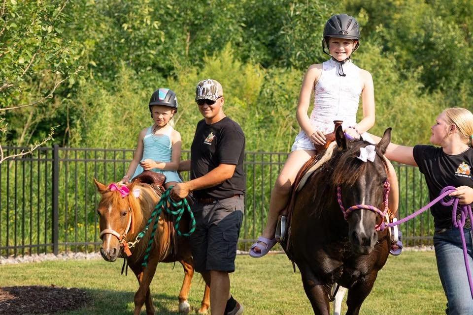 Two girls wearing riding helmets sit on ponies led by two staff members inside a fenced backyard.