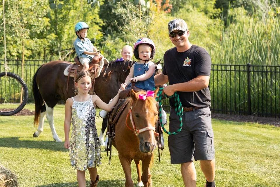 A staff member leads two ponies carrying young children past other kids in a backyard with a tire swing hanging from a tree.