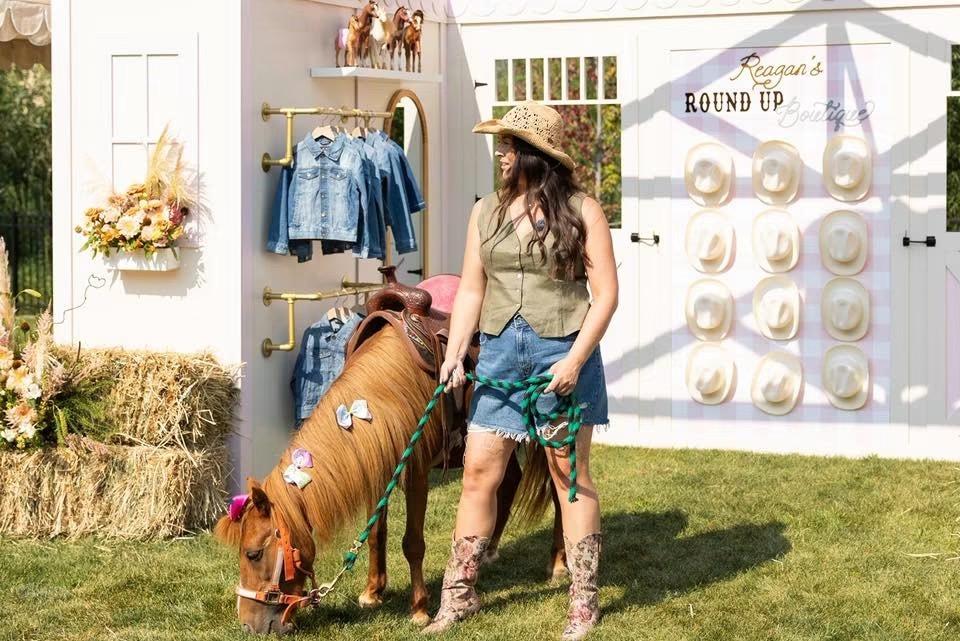 A woman in a western vest and denim skirt leads a pony past a display reading 'Reagan's Round Up Boutique' with denim jackets and hats on racks.