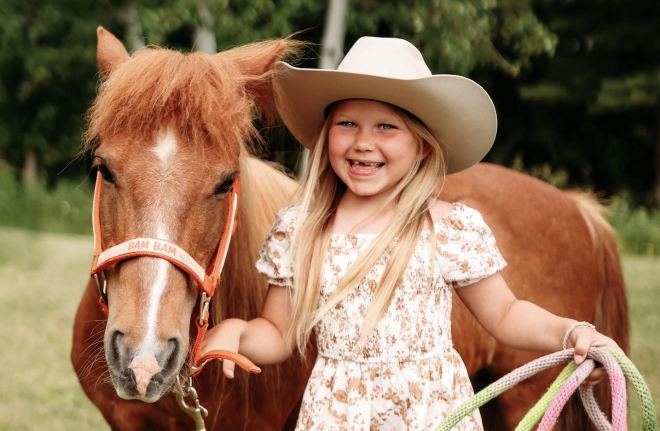 A young cowgirl in a cowboy hat standing with Bam Bam the pony