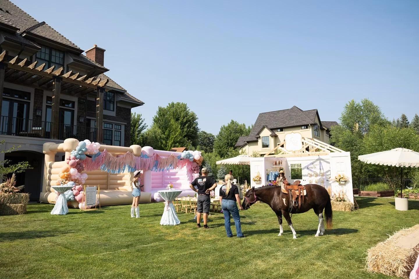 Adults and a saddled horse stand on a lawn near a bounce house, hay bales, and a decorated white booth at a large house.