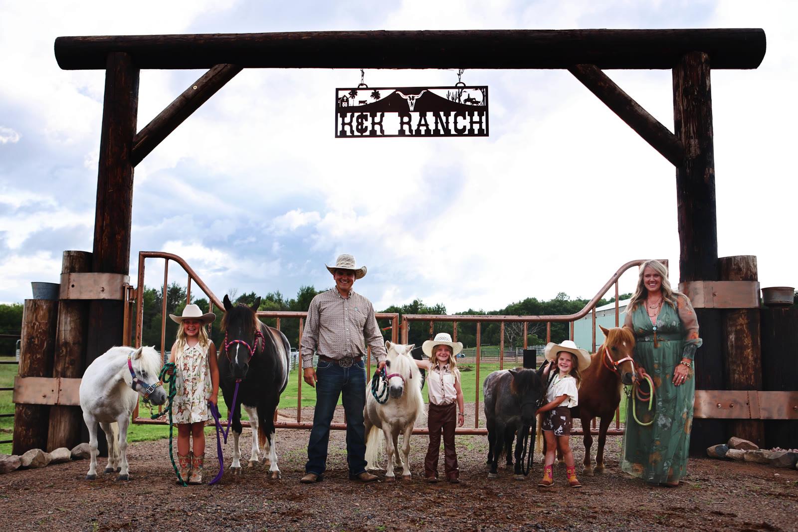 Family of five and two adults pose with five ponies under a wooden ranch entrance sign reading K&K RANCH.