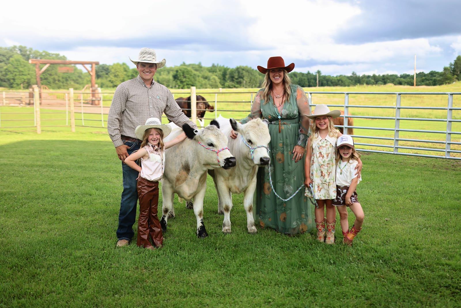 A family of five stands in a pasture with two young cream-colored cattle wearing rope halters.
