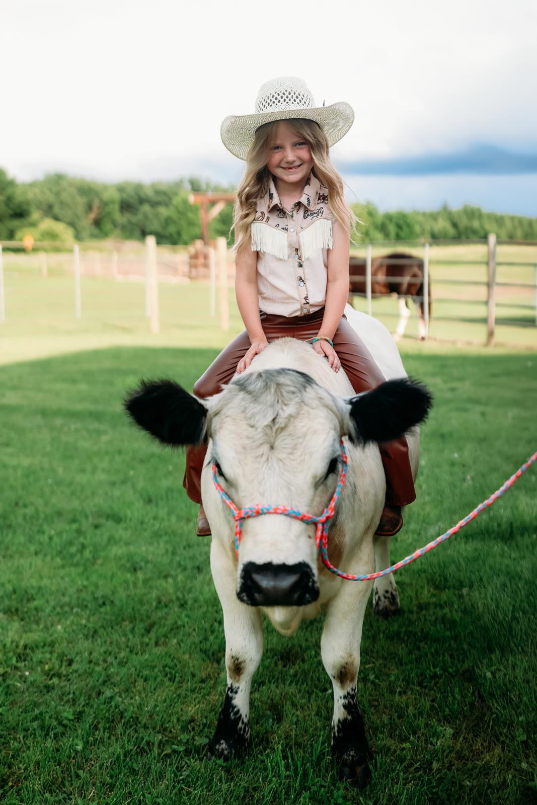 A girl in a cowboy hat sits on the back of a young cream-colored calf in a grassy pasture.