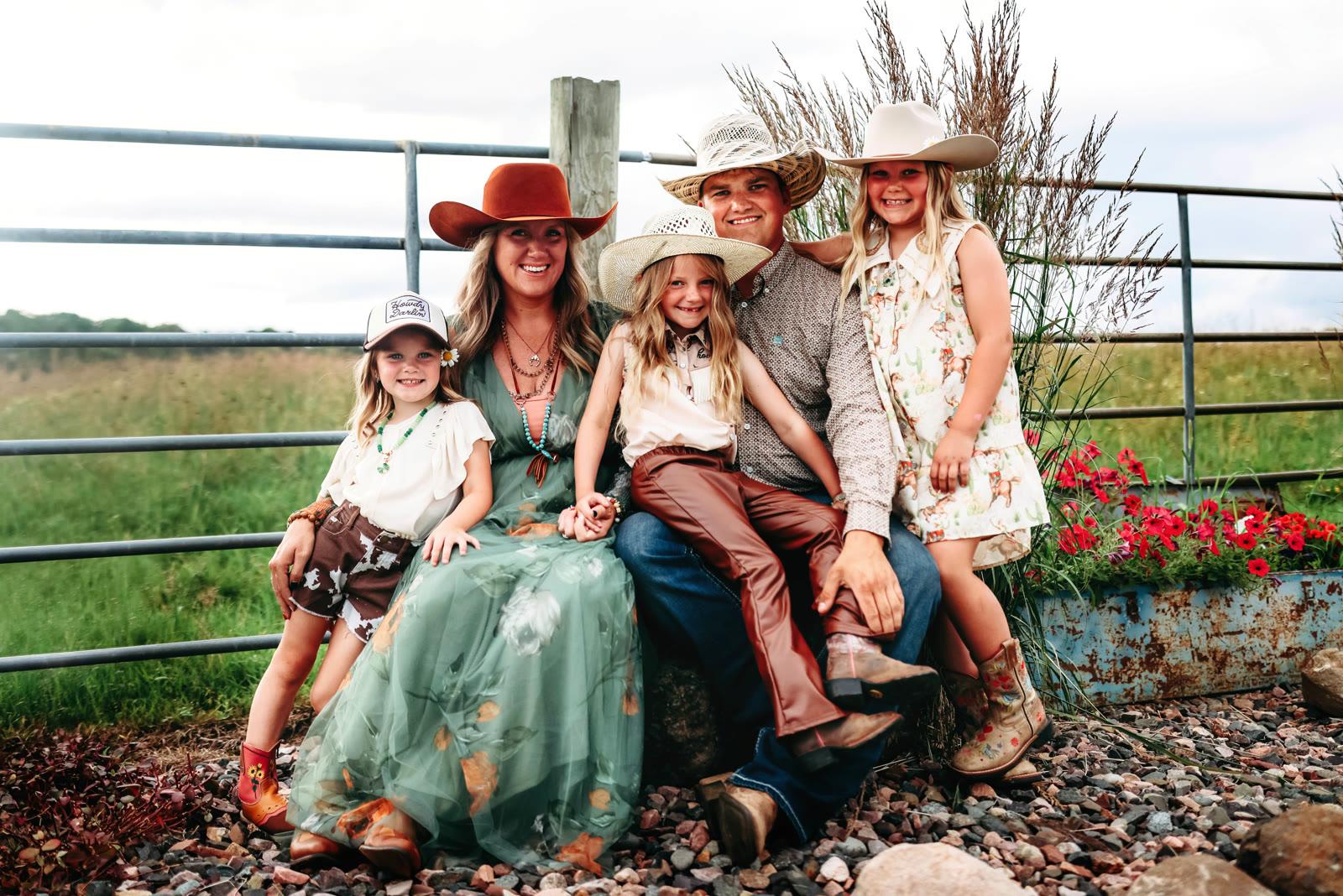 A family of five sits together on a rock wall near a metal gate and red flowers in a grassy field.