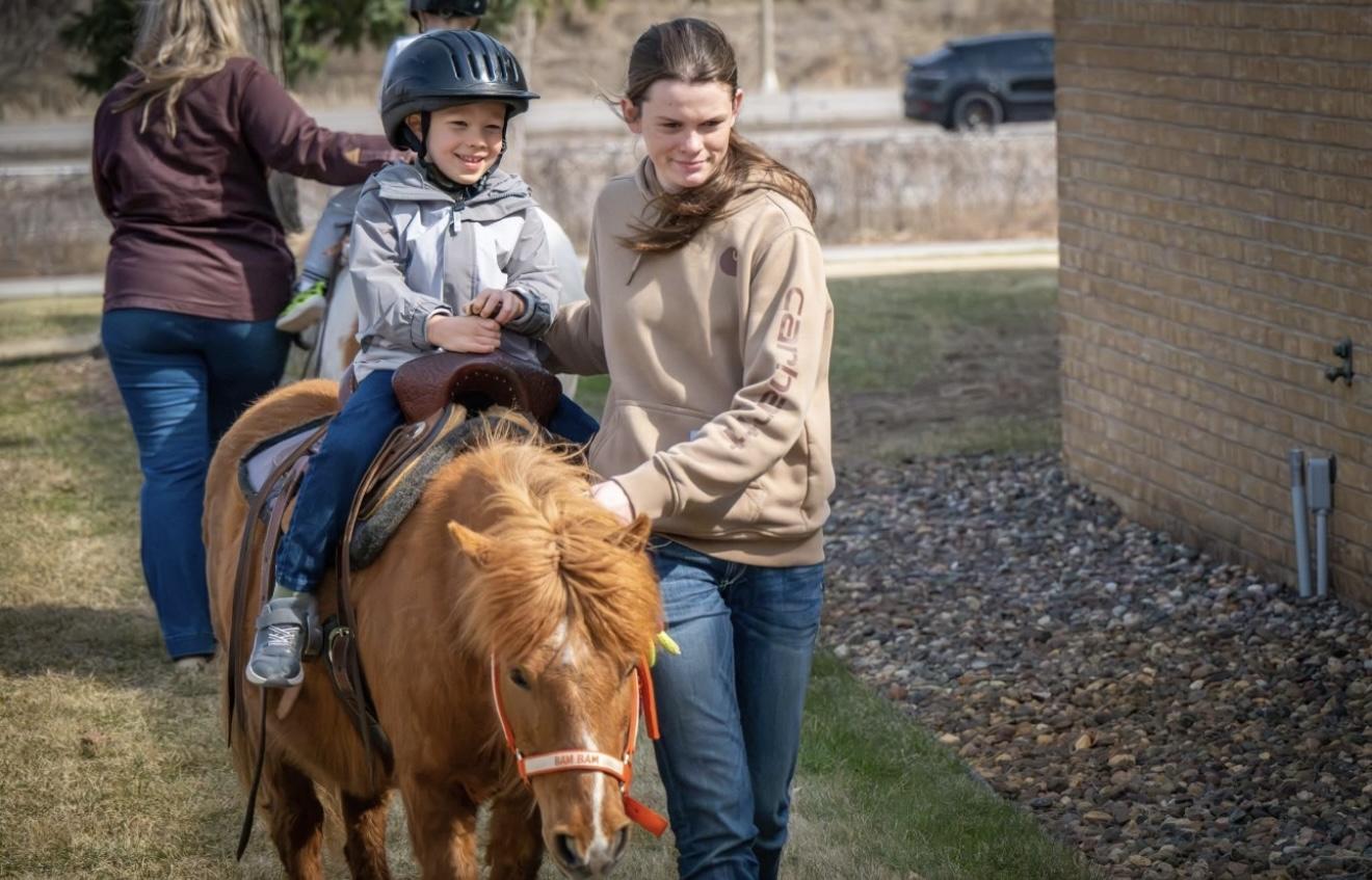 A boy wearing a riding helmet sits on the pony Bam Bam while a handler walks alongside him on a residential driveway.