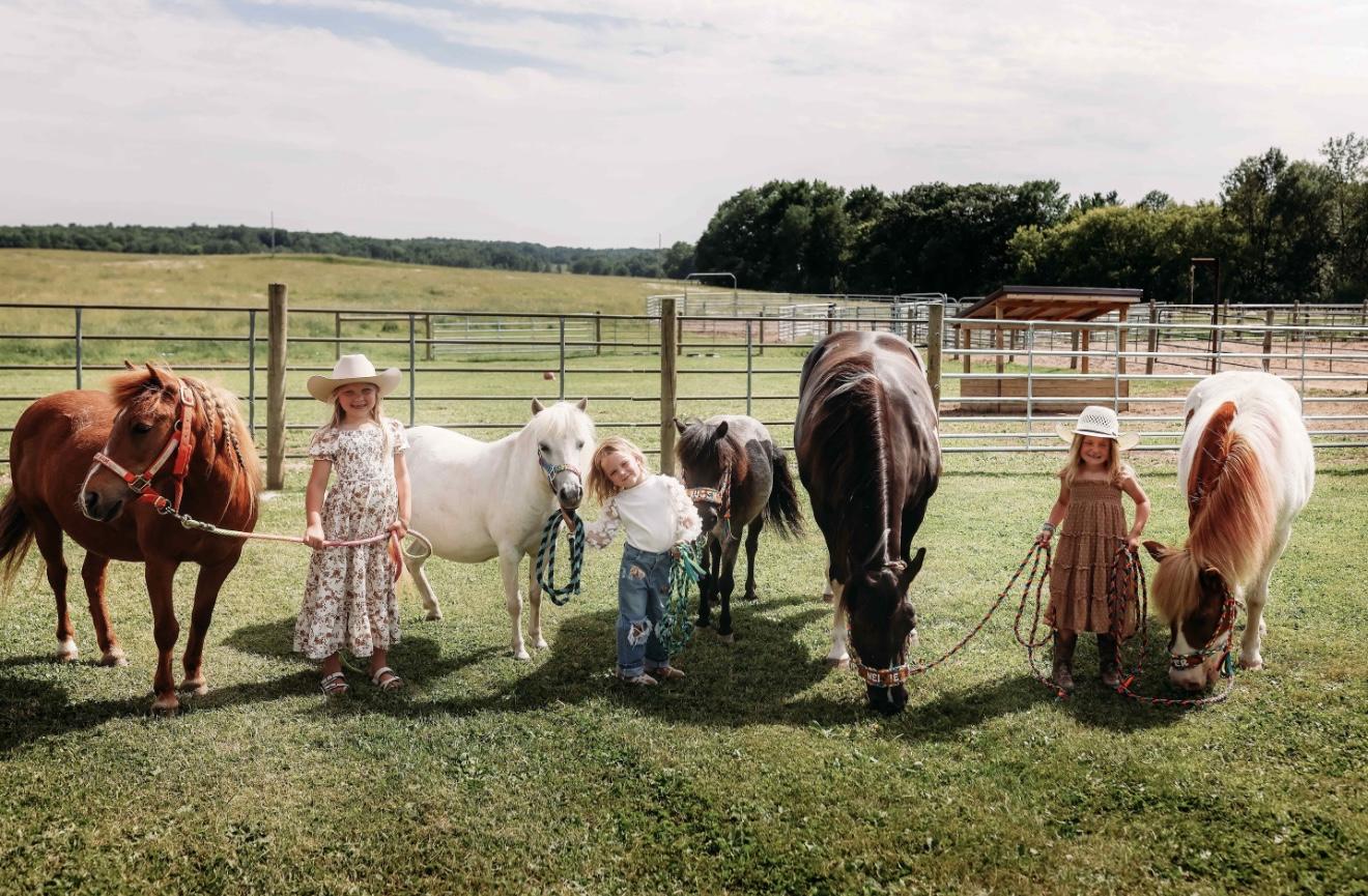 Three girls stand in a fenced pasture holding lead ropes for five ponies and horses of different colors.
