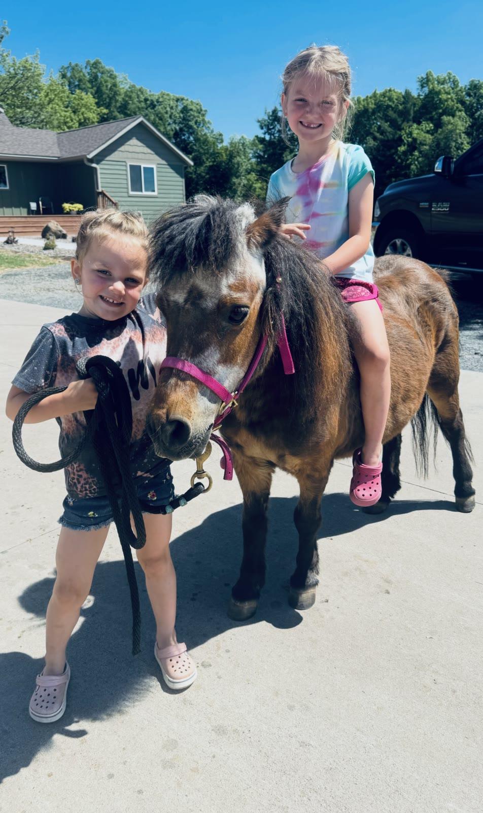 Two girls stand with a pony on a driveway, one sitting on its back, with a house and pickup truck in the background.