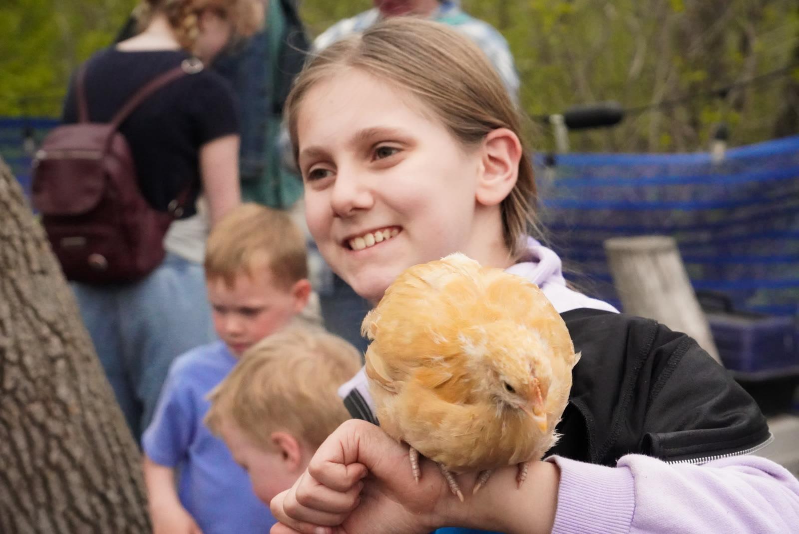 A smiling girl holds a small yellow chick on her arm, with other children nearby outdoors.