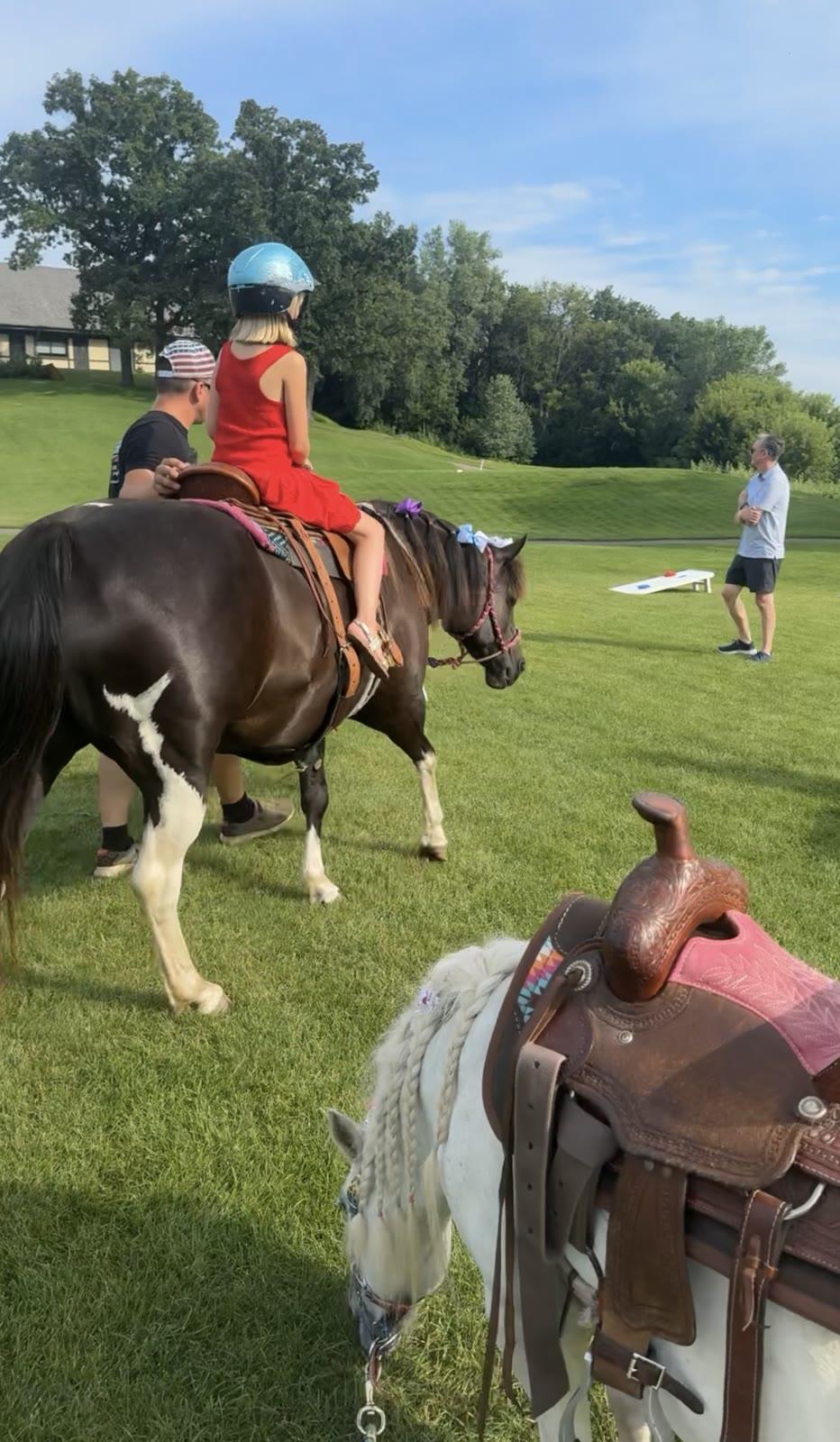A staff member helps fit a helmet on a girl in a red dress next to a pony, with a cornhole board and a man standing in an open lawn area in the background.