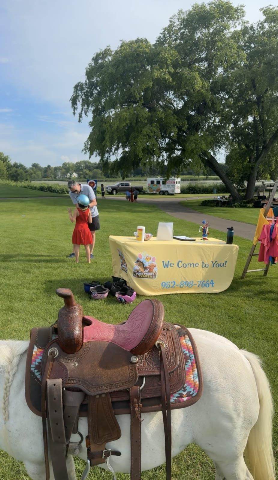 A saddled white pony stands near a yellow table branded 'Pony Parties Express, We Come to You!' with a horse trailer parked on a path in the background.