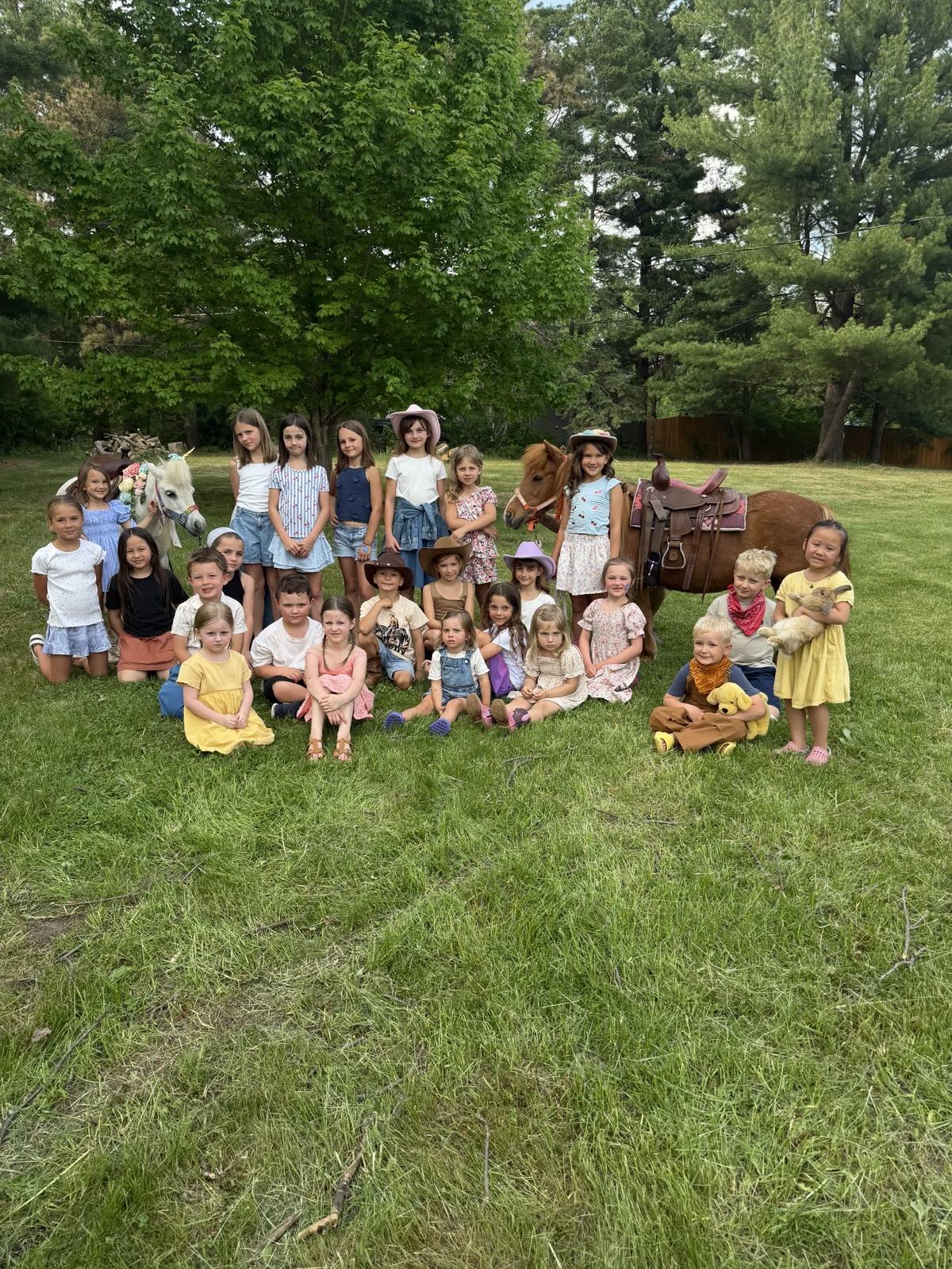 A large group of children pose on a lawn with two ponies, one girl holding a rabbit, in front of a tree line.