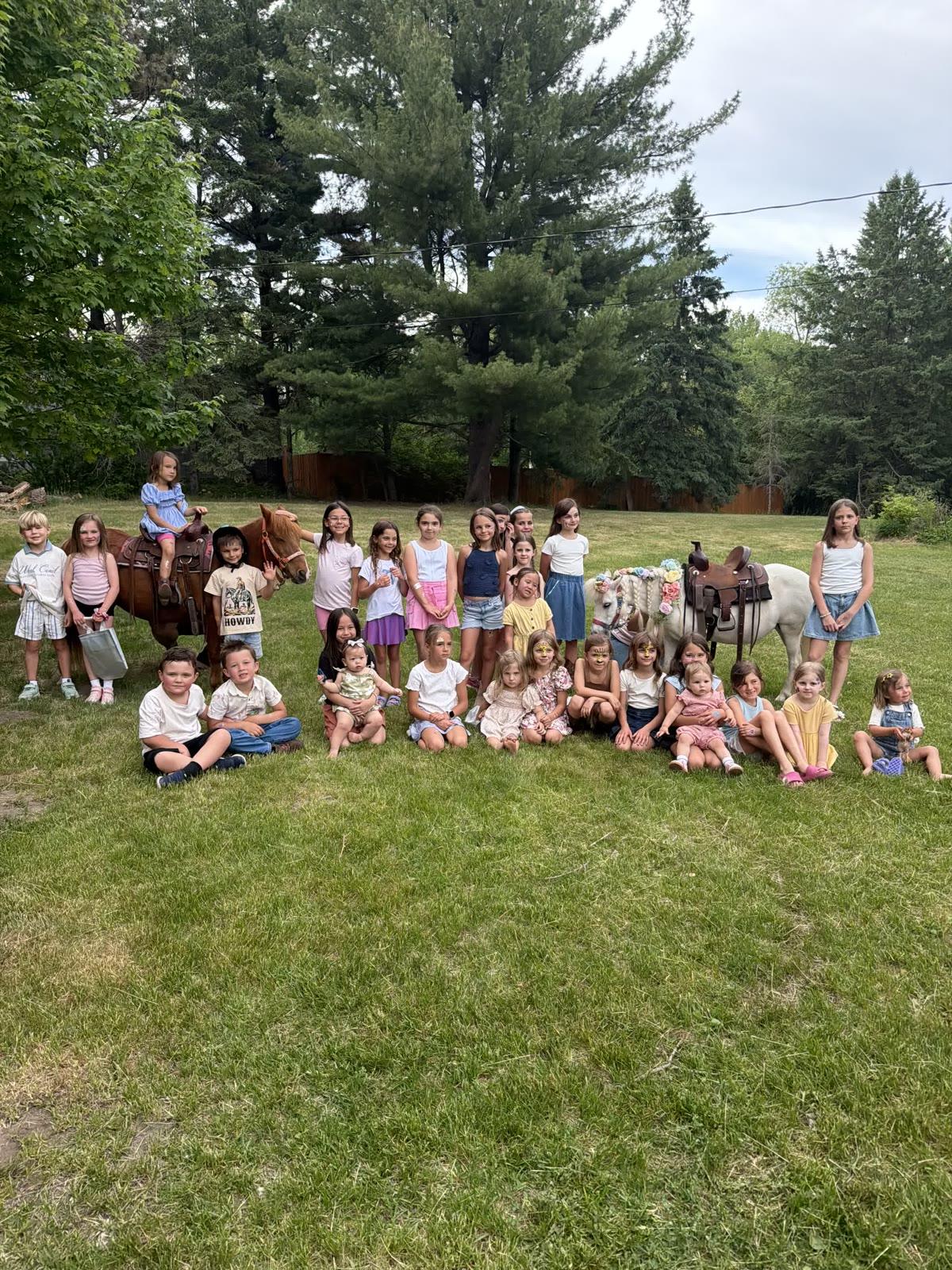 A large group of children pose on a lawn with two ponies near a wooden fence, with one boy wearing a shirt that reads 'Howdy'.