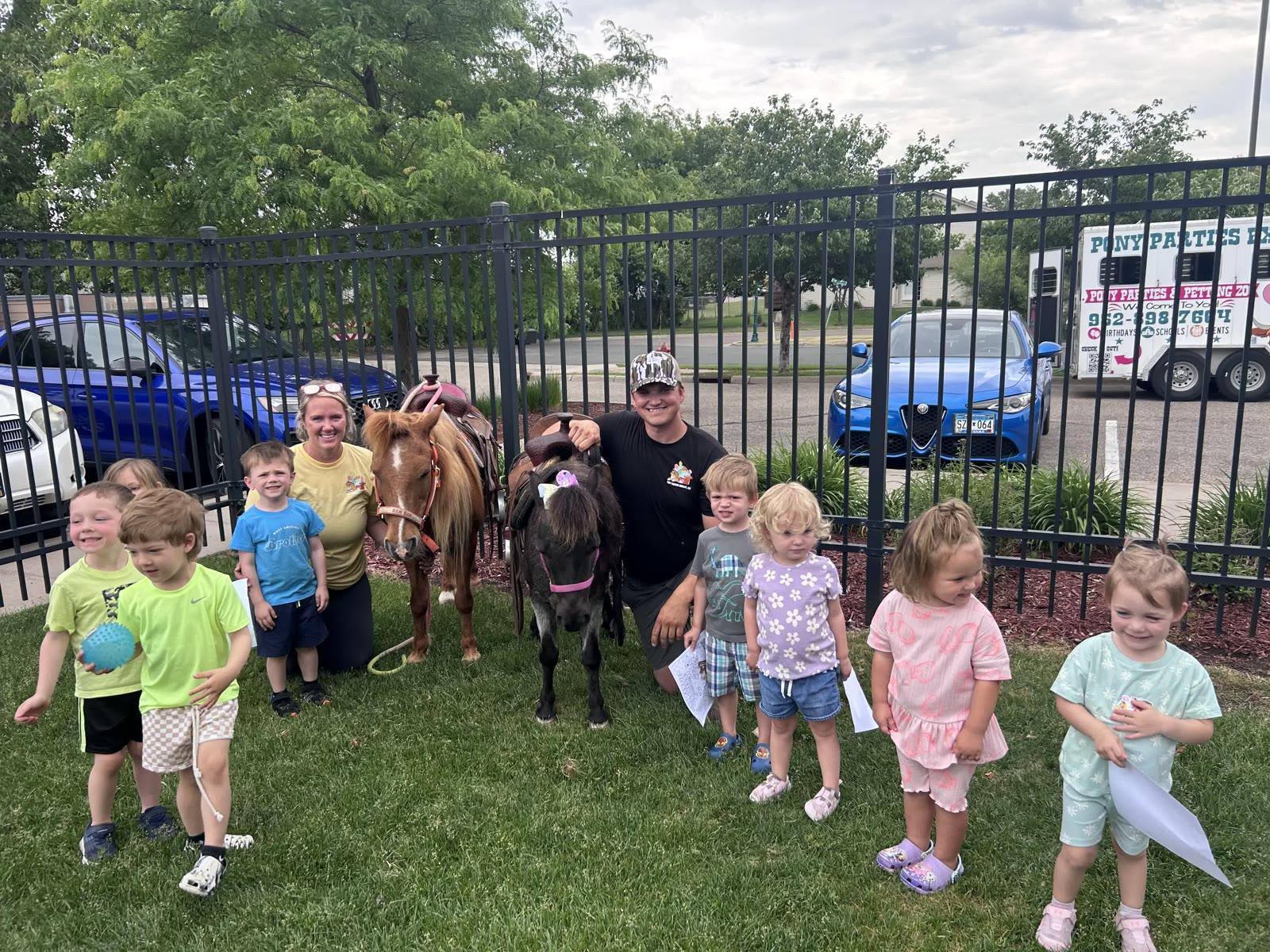 A group of young children stand near two staff members holding two ponies inside a fenced area, with a 'Pony Parties Express' trailer visible through the fence.