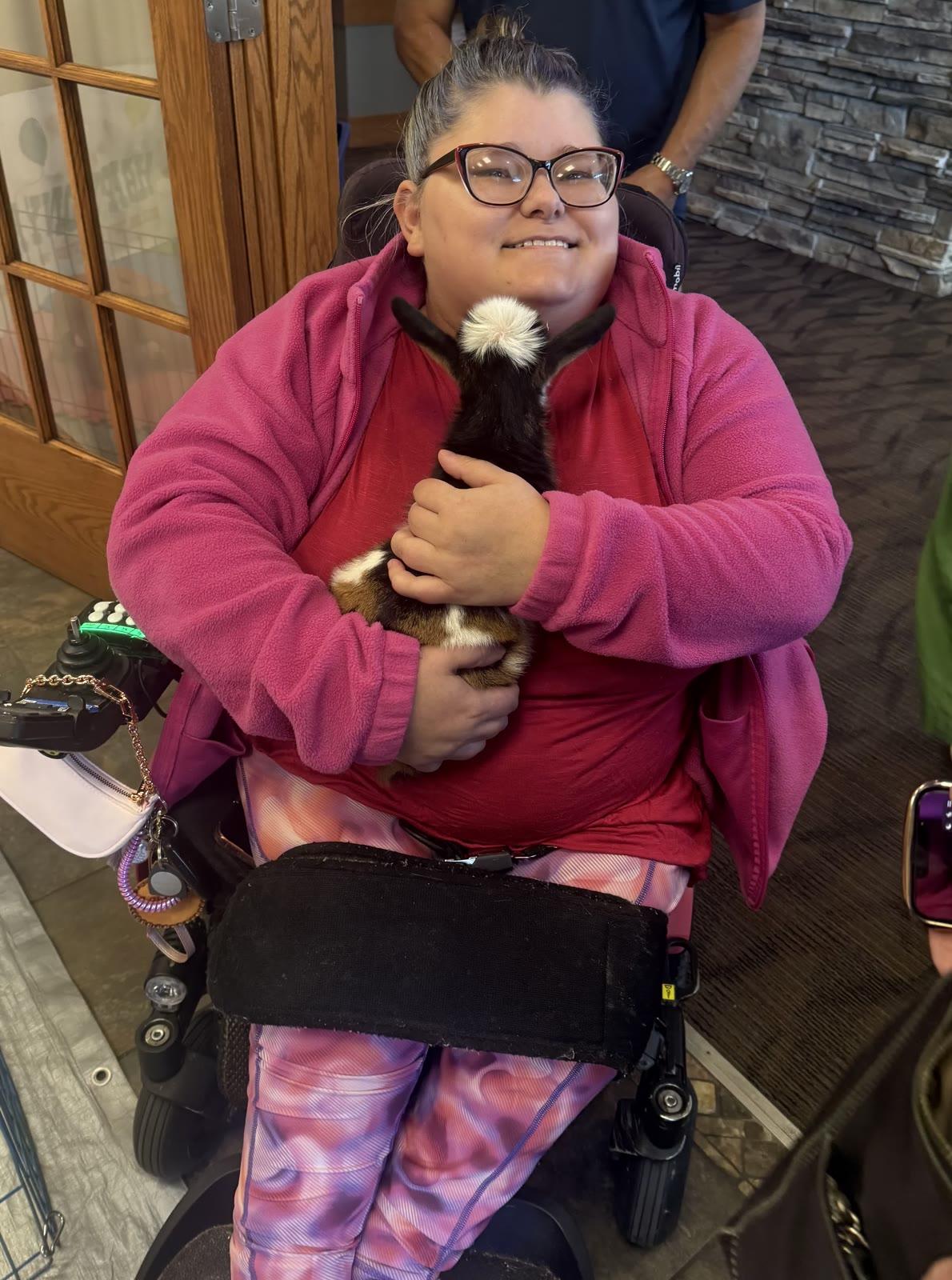 A woman seated in a motorized wheelchair smiles while holding a small black-and-white baby goat, indoors near a stone fireplace.