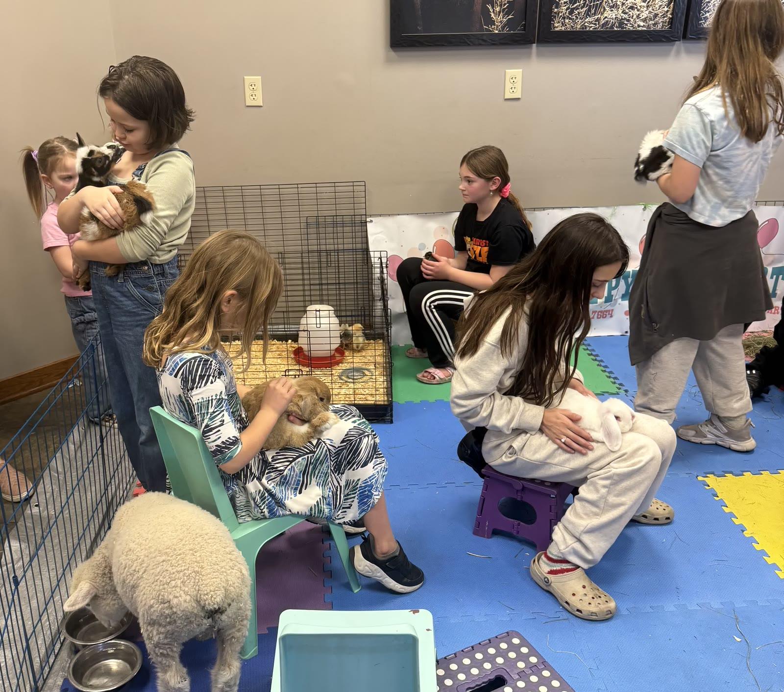 Several children sit and stand indoors on foam floor mats petting rabbits, a lamb, and baby chicks in a wire pen, with a 'Pony Parties Express' banner visible in the background.