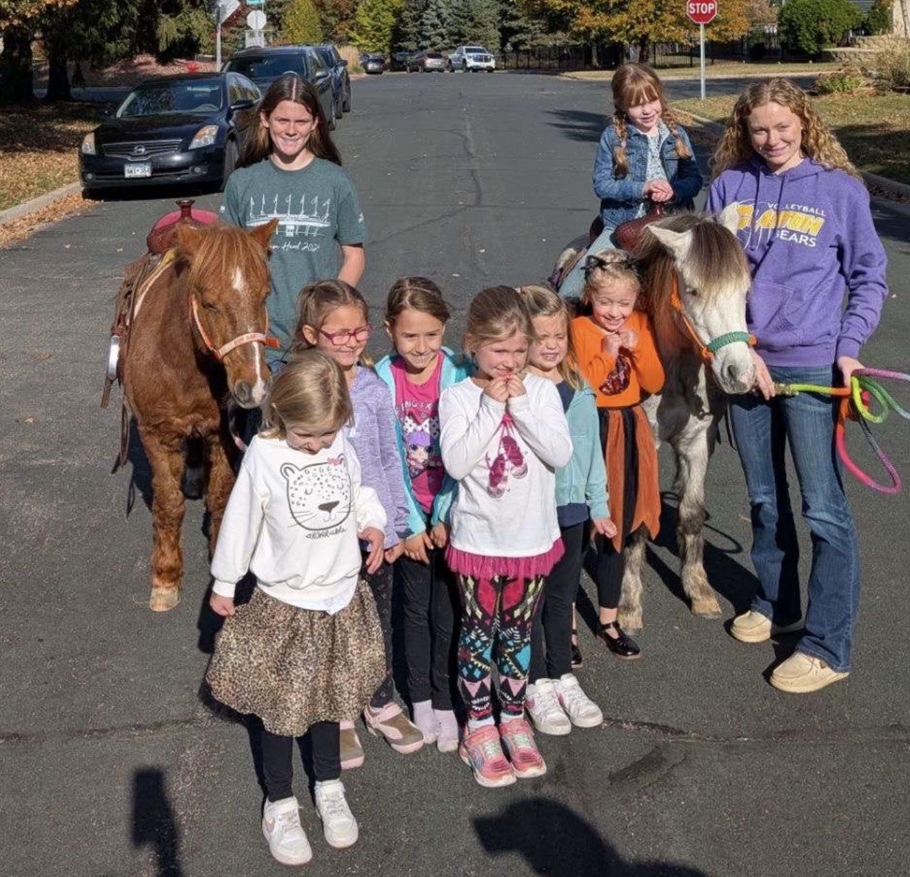 A group of children and two adults stand with two ponies on a residential street.