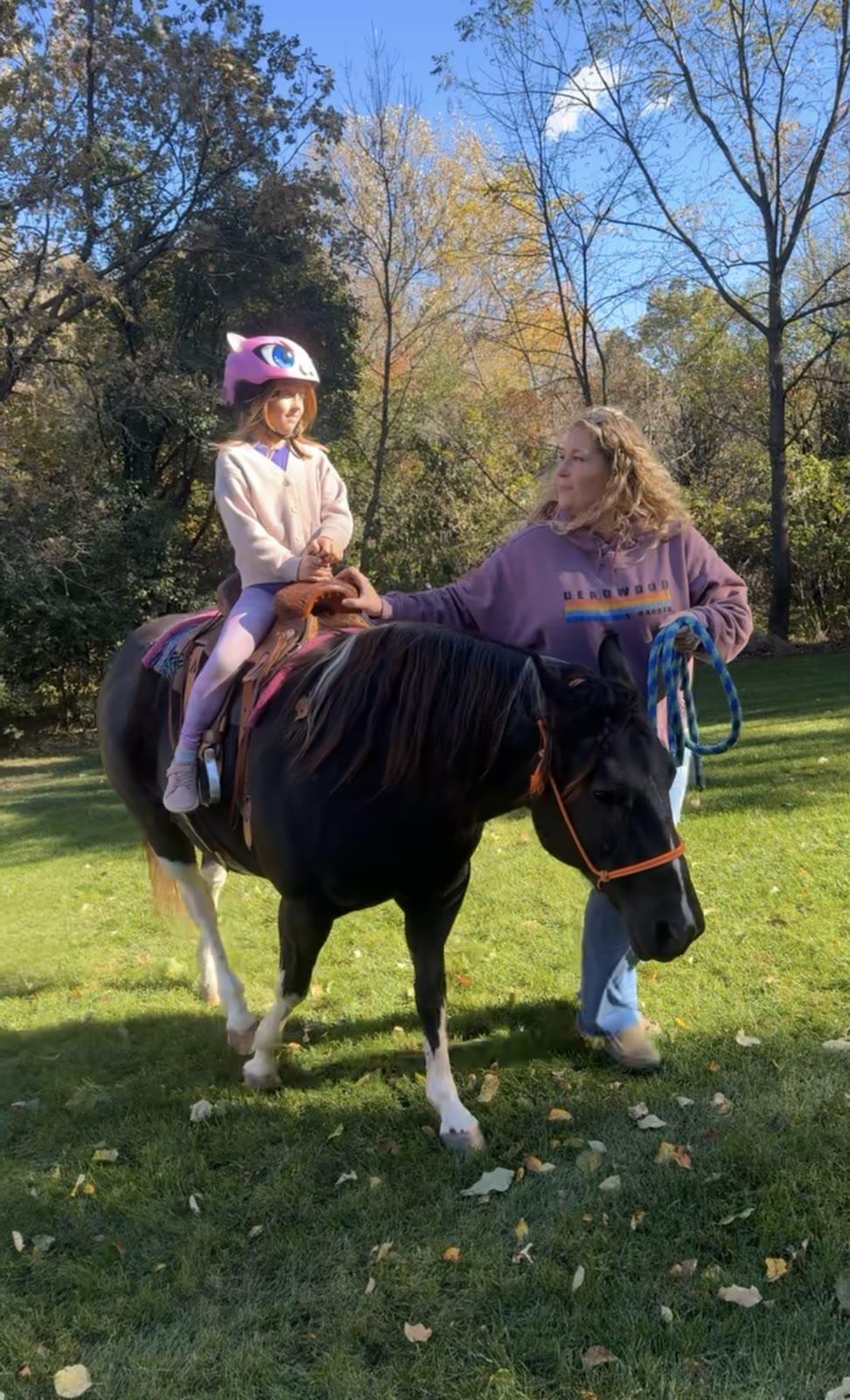 A child wearing a unicorn helmet sits on a black-and-white pony while a woman in a purple hoodie holds the lead in a grassy yard.