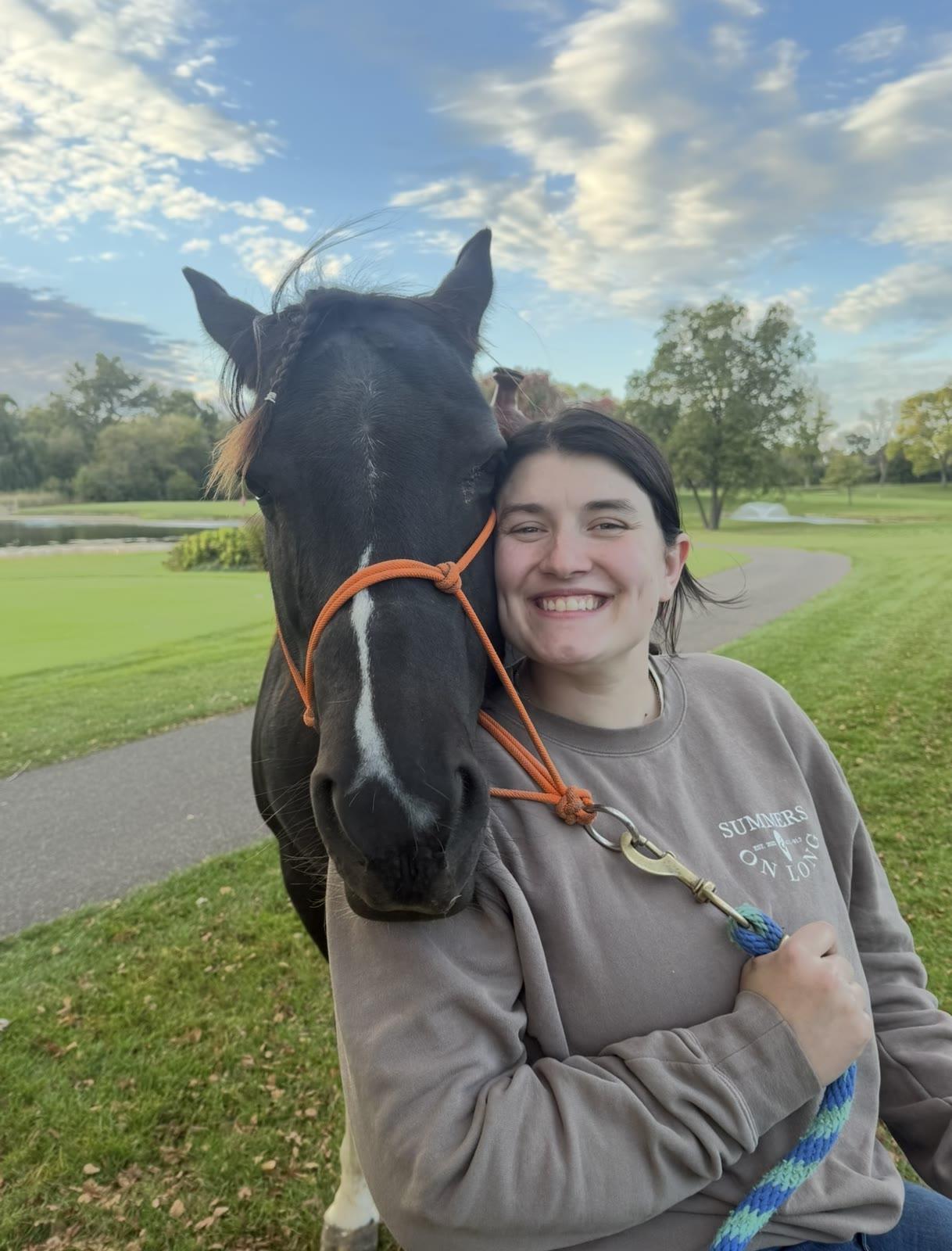 A woman hugs the head of a black horse on a grassy path near a pond.