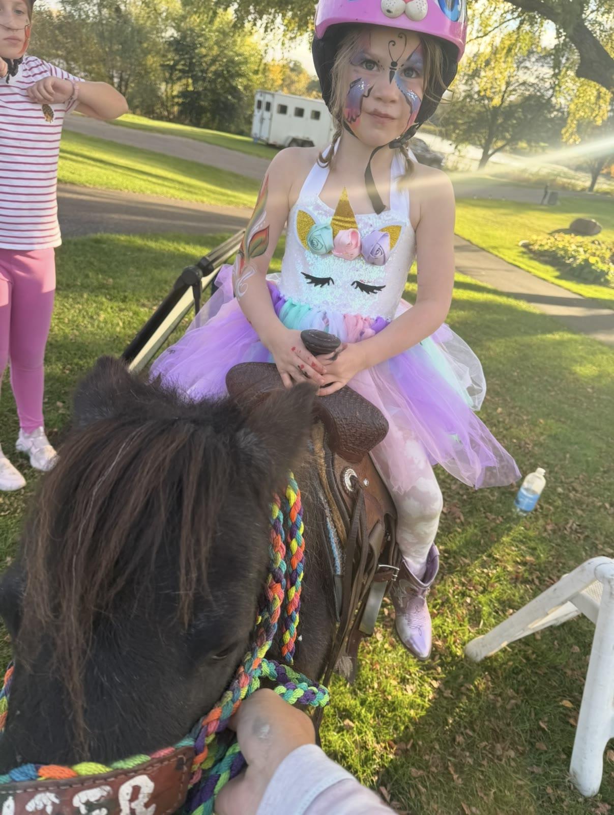 A child in a unicorn costume with face paint sits in a saddle on a pony, with a horse trailer in the background.