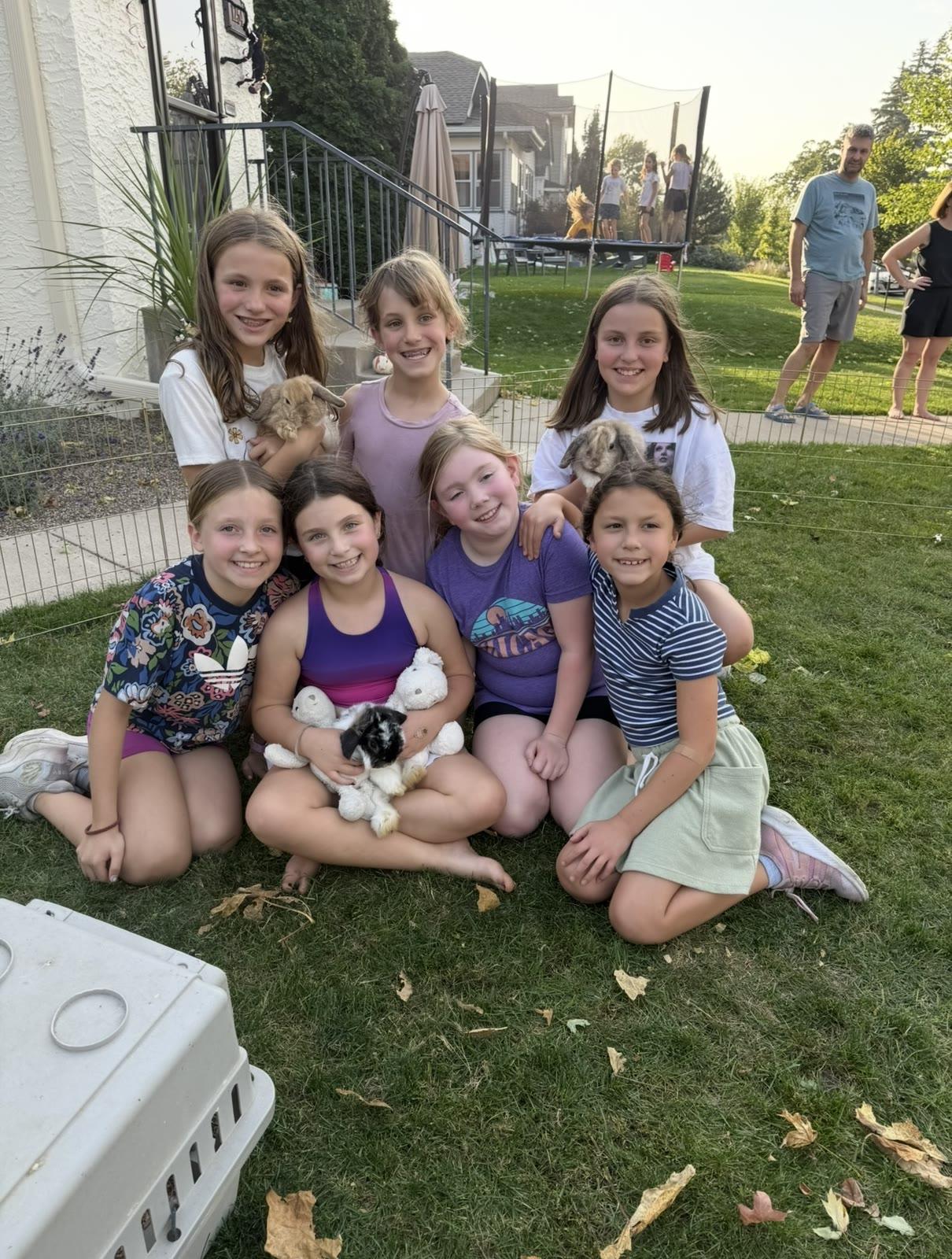 A group of children kneel together on grass holding two rabbits in a backyard with a trampoline.