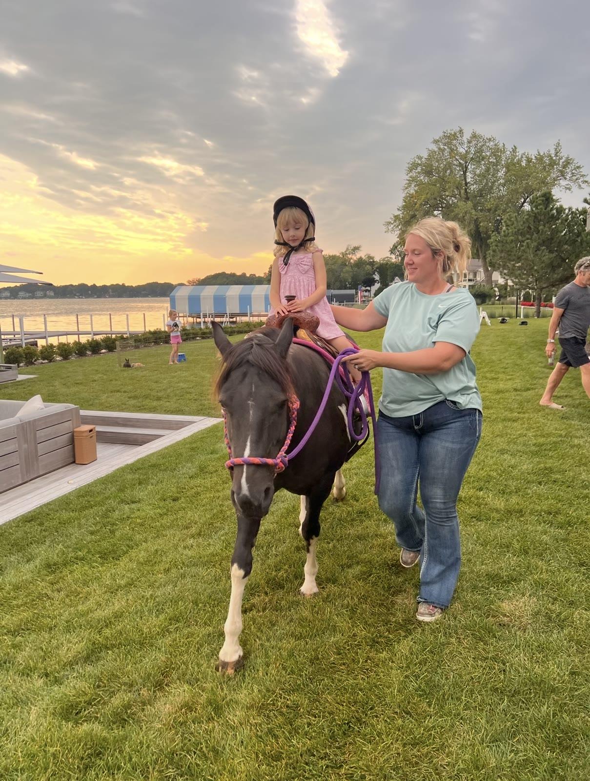 A child in a helmet sits on a pony led by a woman on a lawn near a lake at sunset.