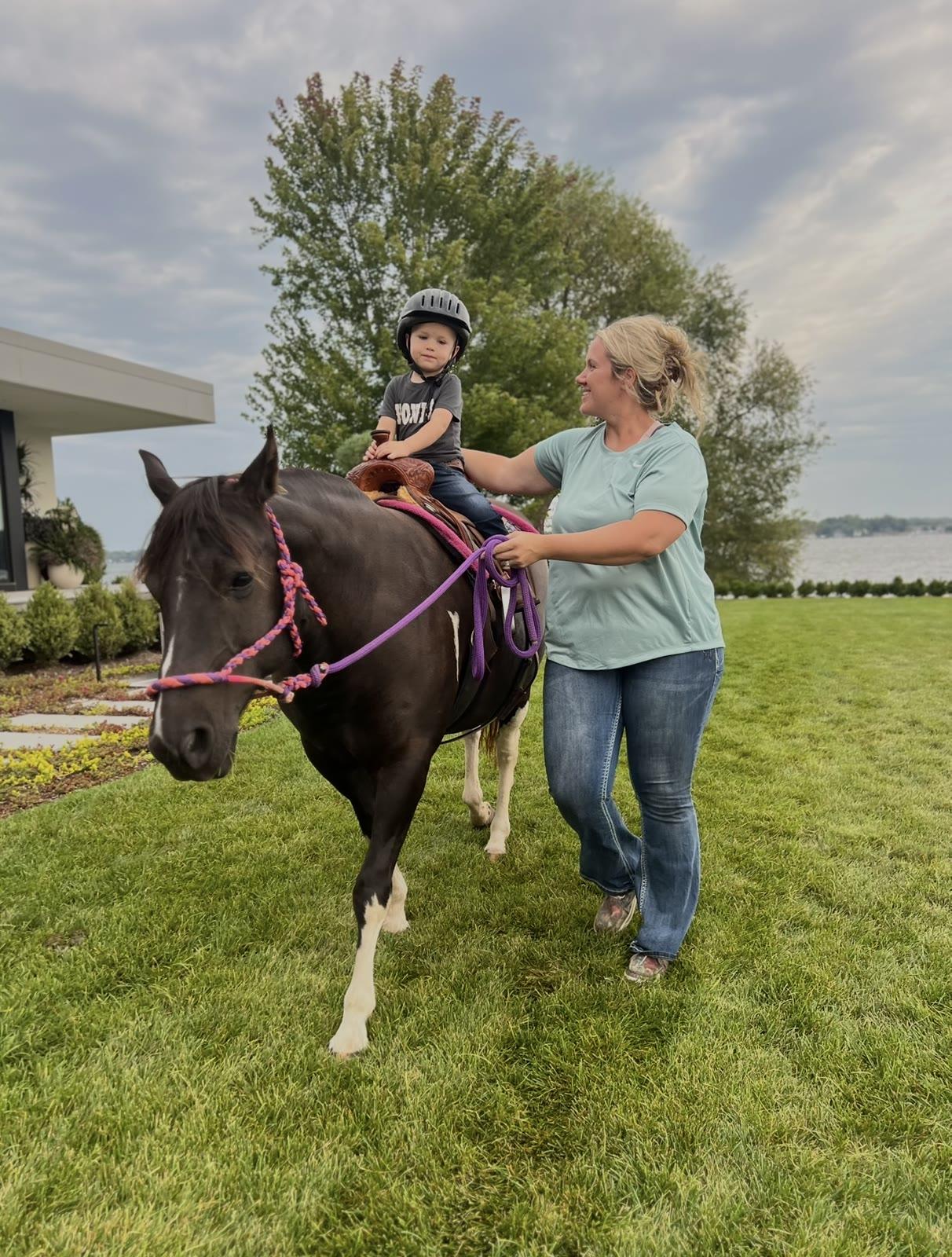 A toddler in a helmet sits on a pony led by a woman on a lawn near a lake.