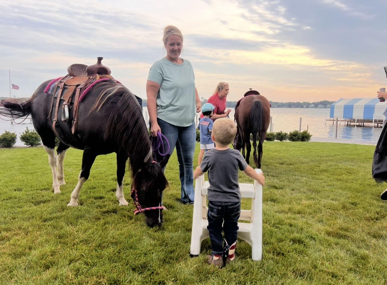 A woman leads a pony across a lawn while a boy stands on a step stool waiting, with a lake and dock in the background.