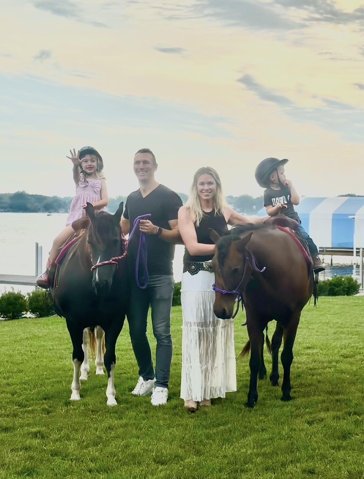 A family of four stands with two children on ponies on a lawn near a lake at sunset.