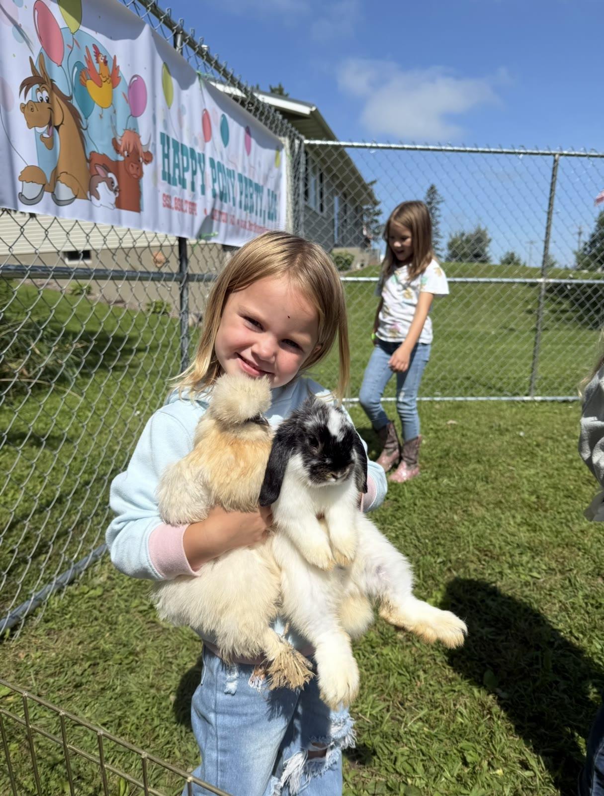 A child holds two rabbits in a fenced pen with a Happy Pony Party banner in the background.