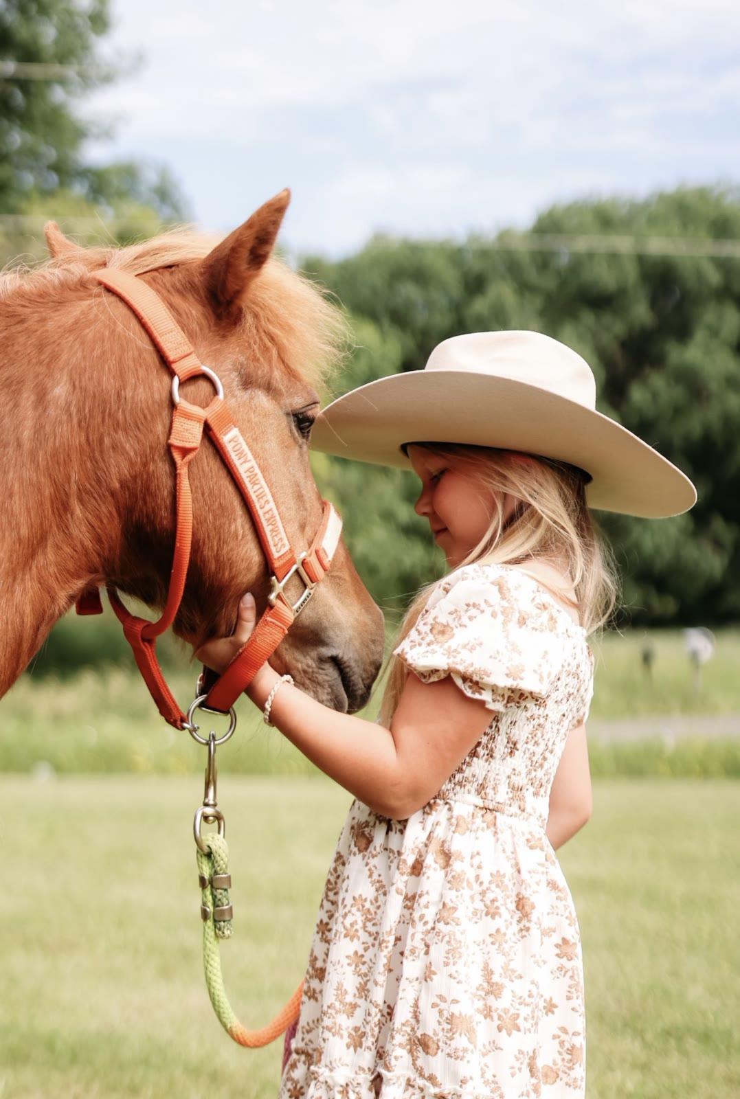 A child in a cowboy hat pets the face of a brown pony wearing a halter labeled Pony Parties Express in a field.