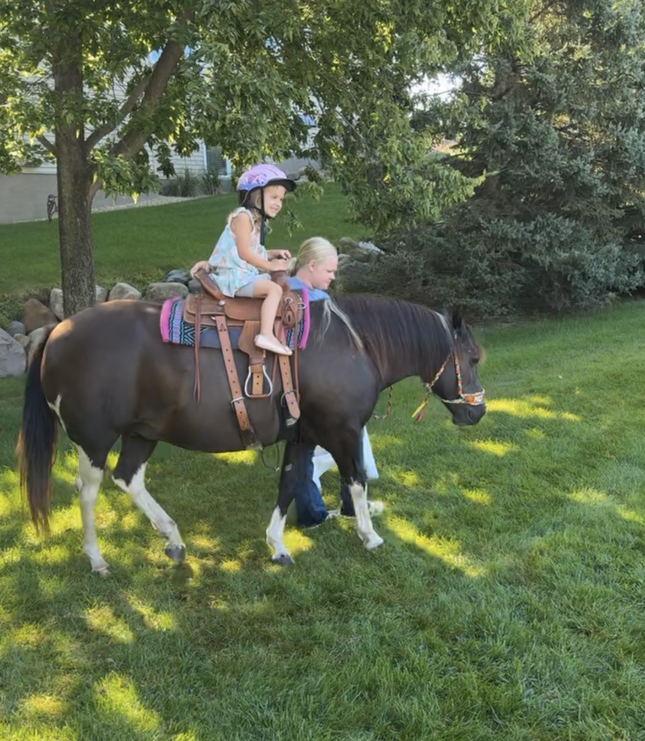 A child in a pink helmet rides a dark pony led by another girl through a shaded backyard.