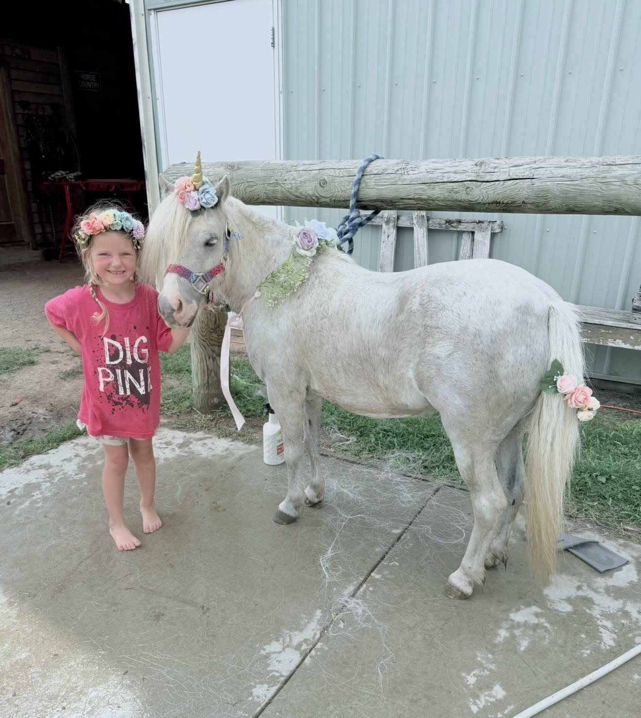 A child wearing a flower crown stands beside a white pony decorated with a unicorn horn and flowers near a barn.