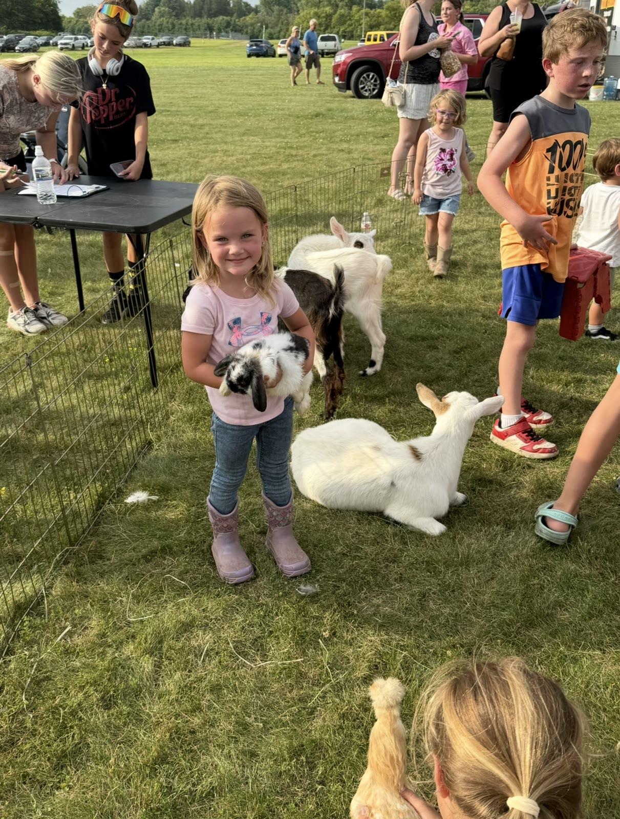 A child holds a black-and-white rabbit near baby goats in a fenced pen at an outdoor event.