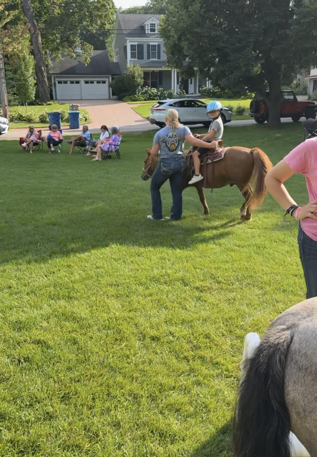 A woman leads a pony with a child rider across a lawn while seated adults watch from lawn chairs near a house.