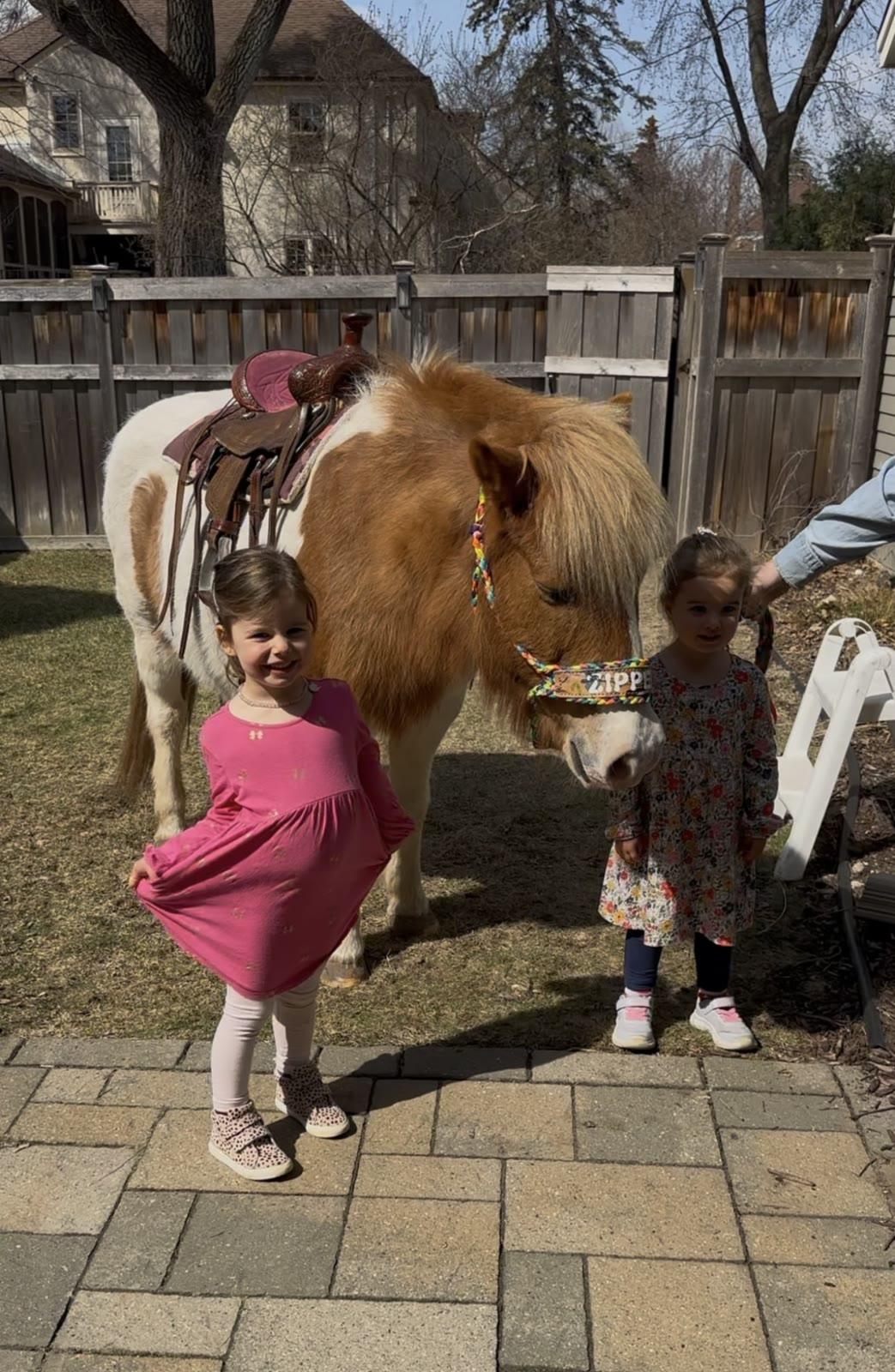Two children stand beside a saddled brown and white pony named Zipper in a backyard.